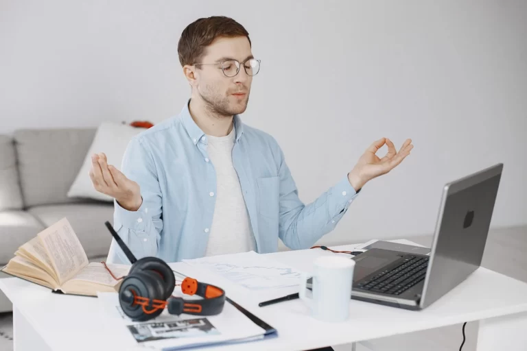 Man meditating at desk with laptop, headset, and documents in home office.