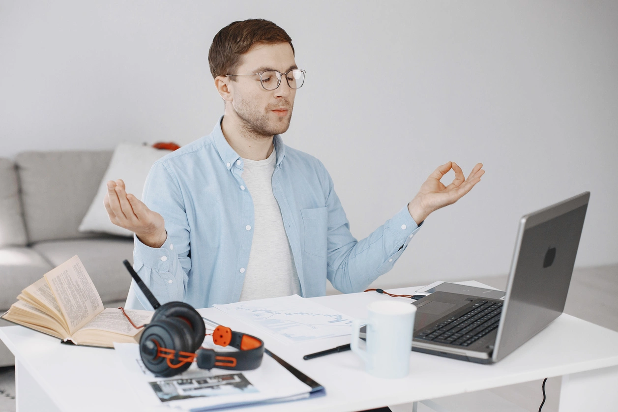 Man meditating at desk with laptop, headset, and documents in home office.